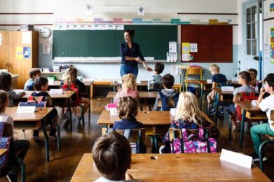 Photo couleur d'une salle de classe avec des enfants aux pupitres. Au fond vers le tableau, une enseignante en action.